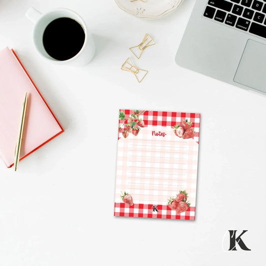 Red gingham notepad with strawberries on desk, beside coffee, pen, and laptop.